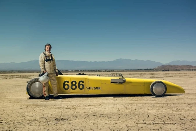 Man in racing suit stands with a yellow land speed record car on cracked desert, with mountains and blue sky in the distance.